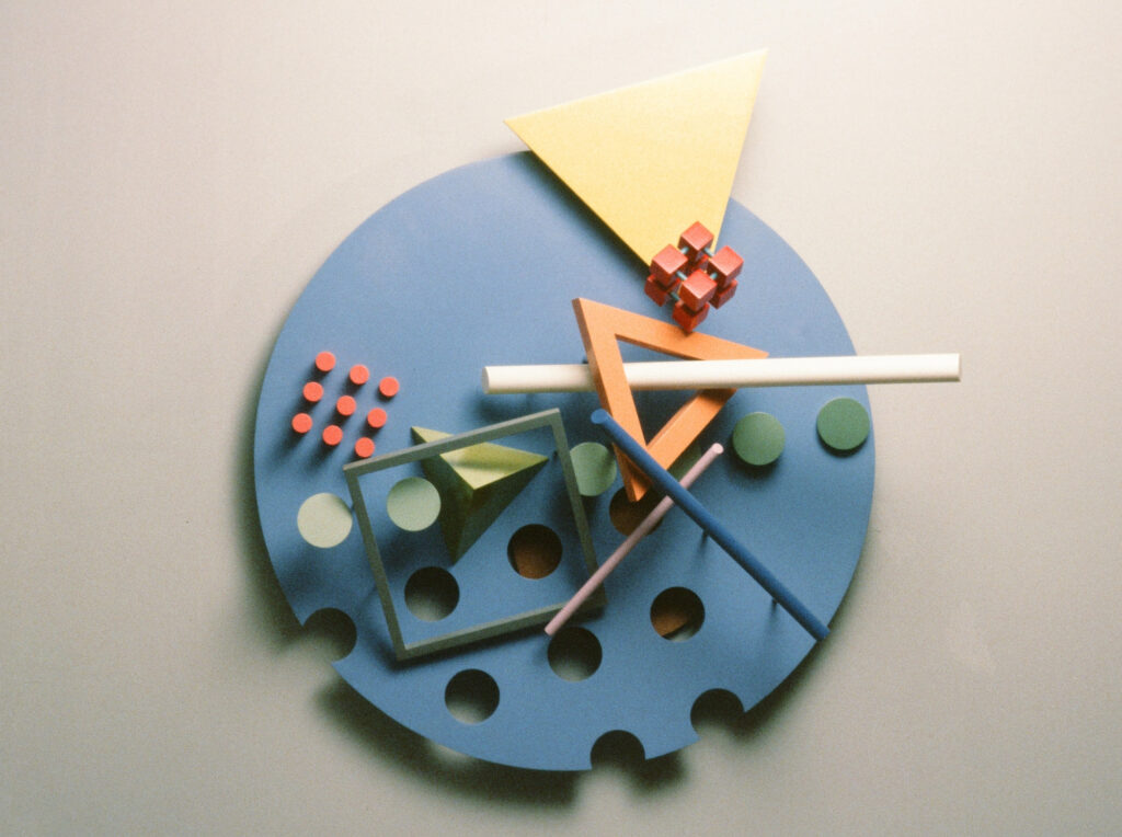 1985 wall sculpture titled 'Fast Walker' featuring a blue circular base with geometric cutouts and various colorful 3D shapes including a yellow triangle, red cubes, orange open triangle, green circles, and intersecting rods